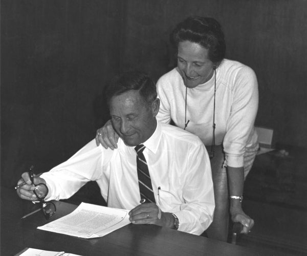 Vieve Gore standing behind her husband, Bill Gore, who is signing a document.